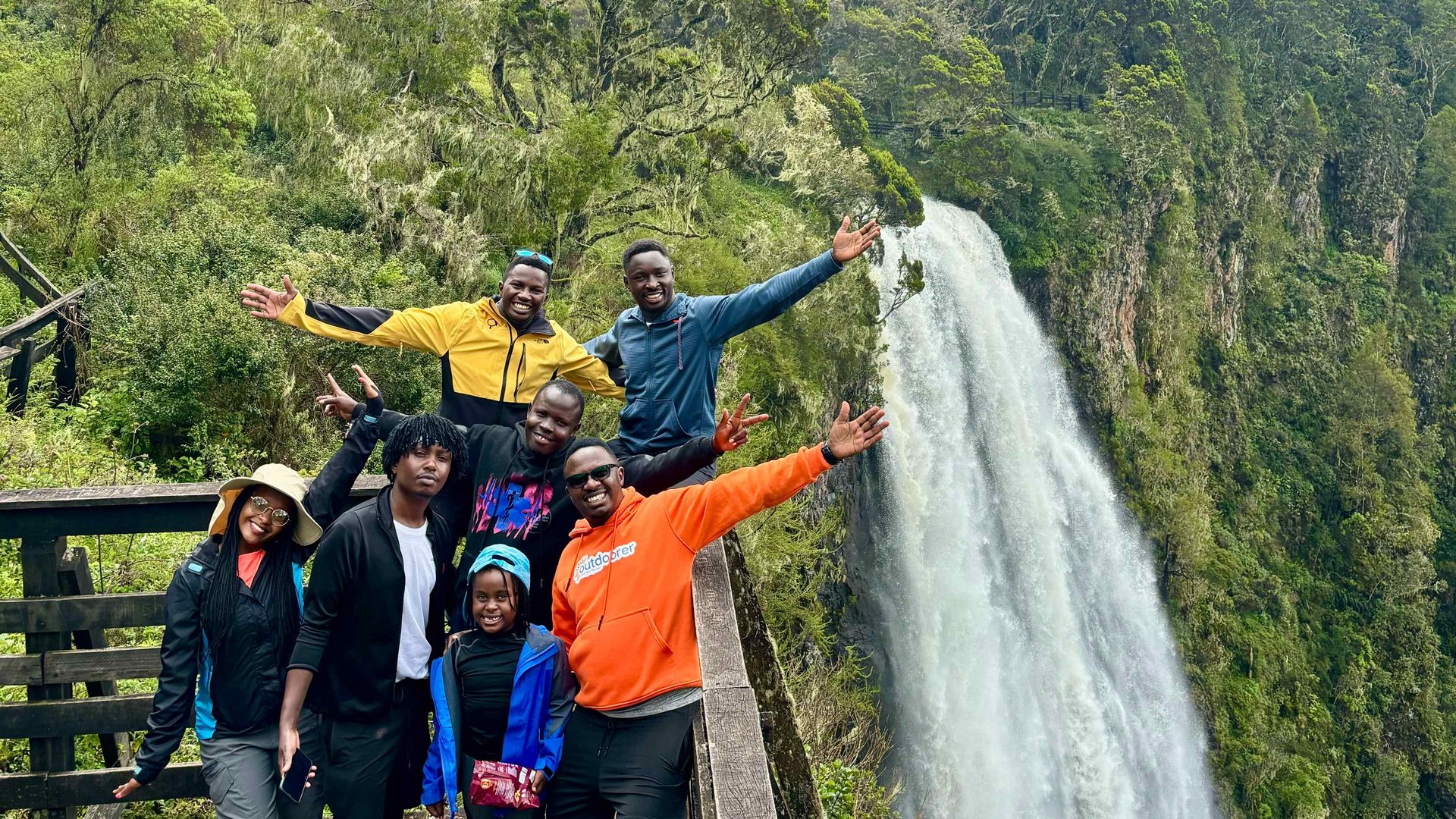 Hikers standing near a ledge posing for a photo in front of a large waterfall | rain for Mt. Kenya, Outdoorer KE