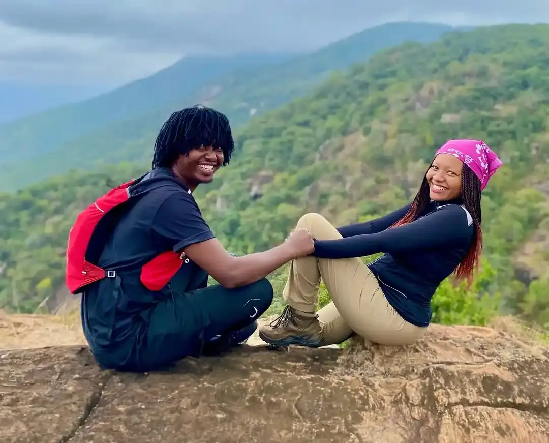 Two hikers sitting on a rock smiling while holding hands, facing the camera. The background shows a scenic mountainerous region.